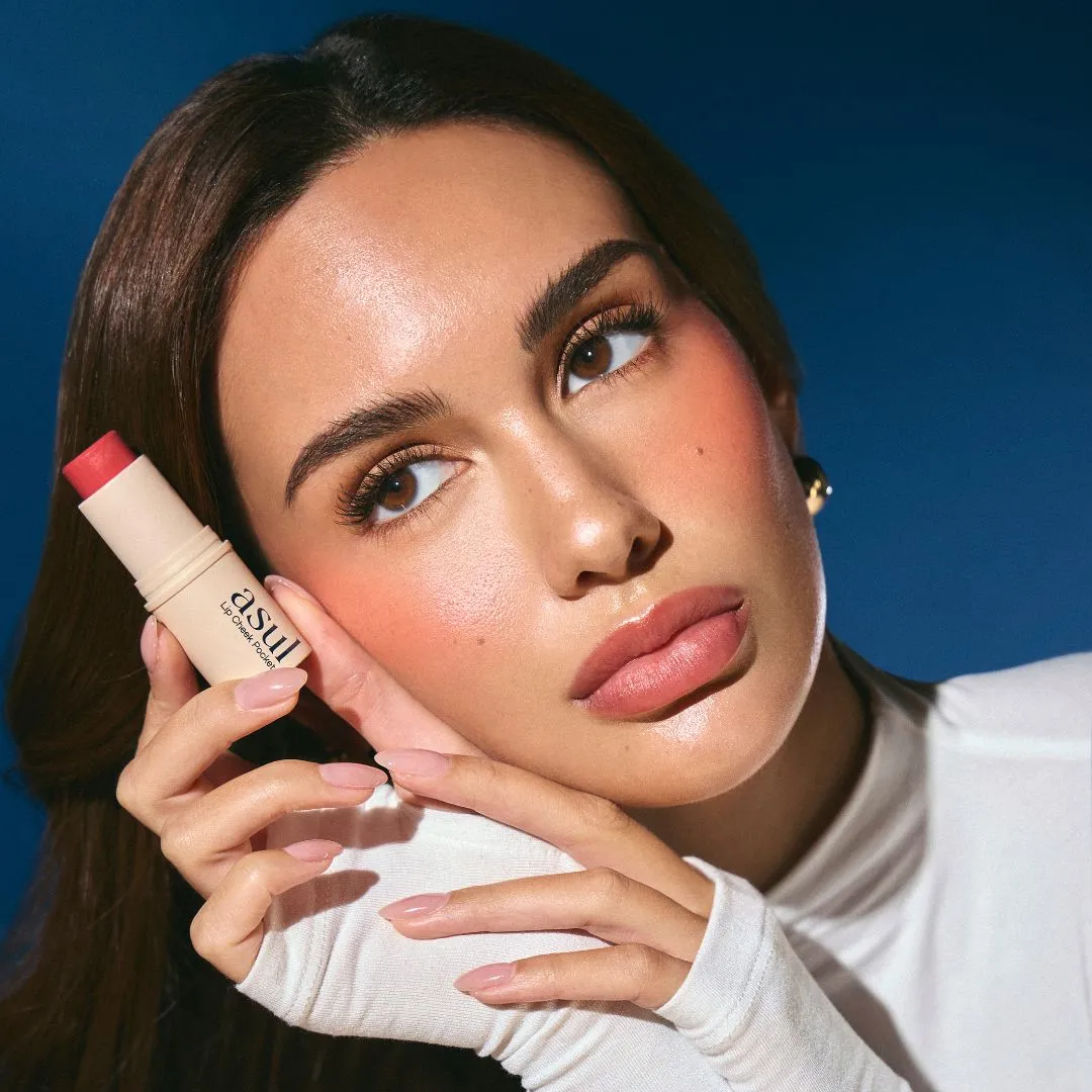 Close-up of a woman with glossy makeup, holding a pink lip balm near her lips against a blue background.