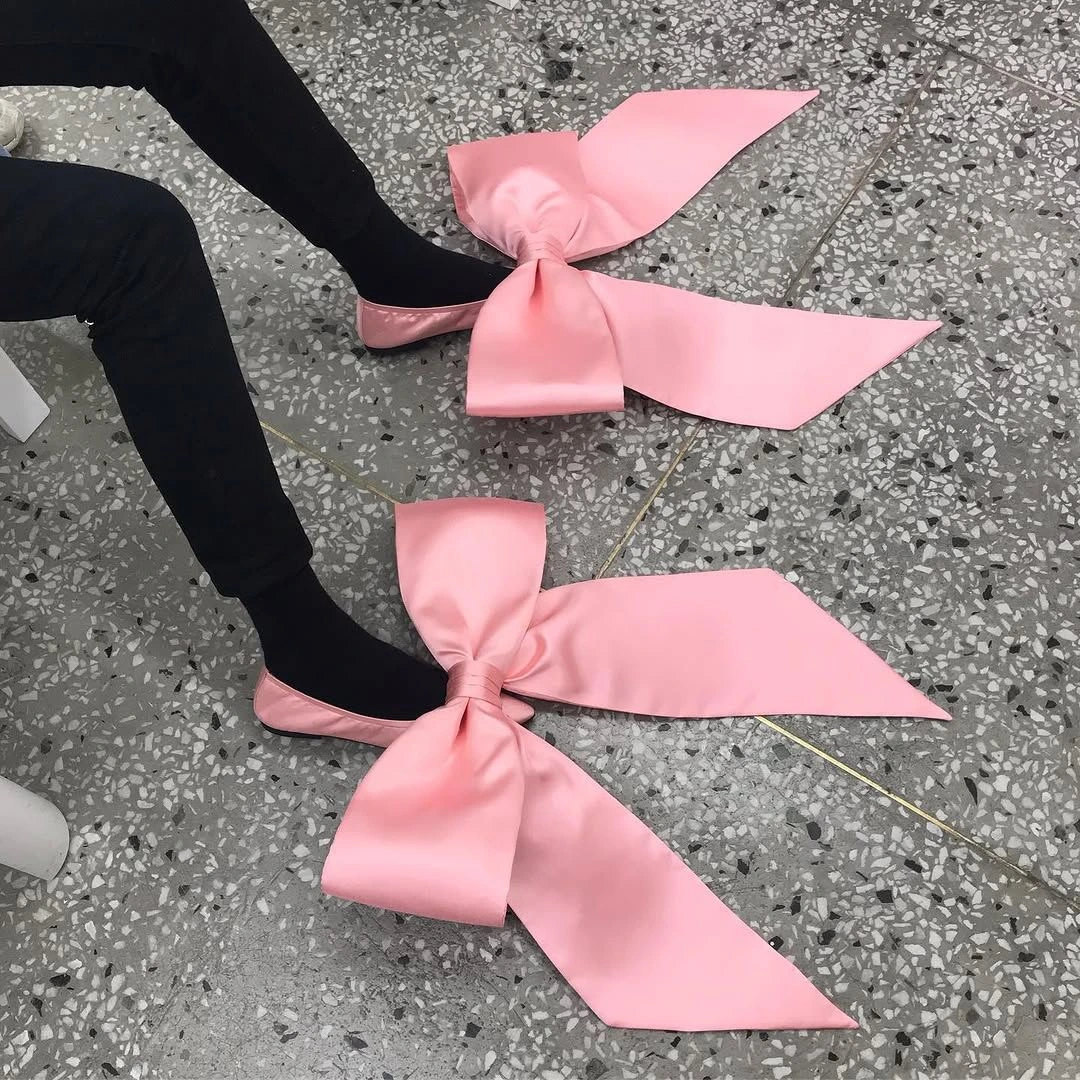 Two large pink satin bow ribbons with long tails lying on a speckled floor beside a person in black socks and pants.
