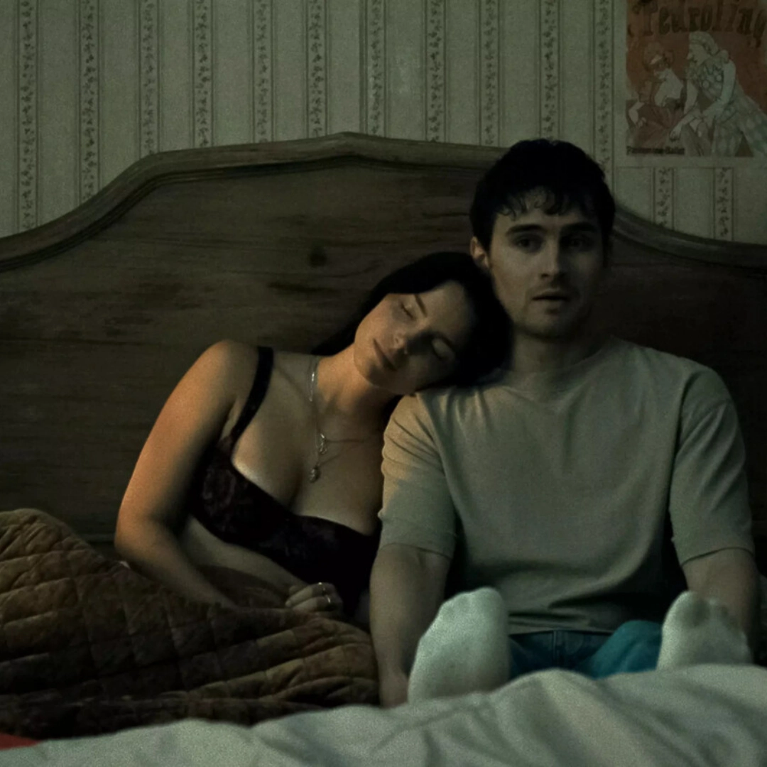Young man sits upright in bed as a woman rests her head on his shoulder, eyes closed, in a dimly lit bedroom.