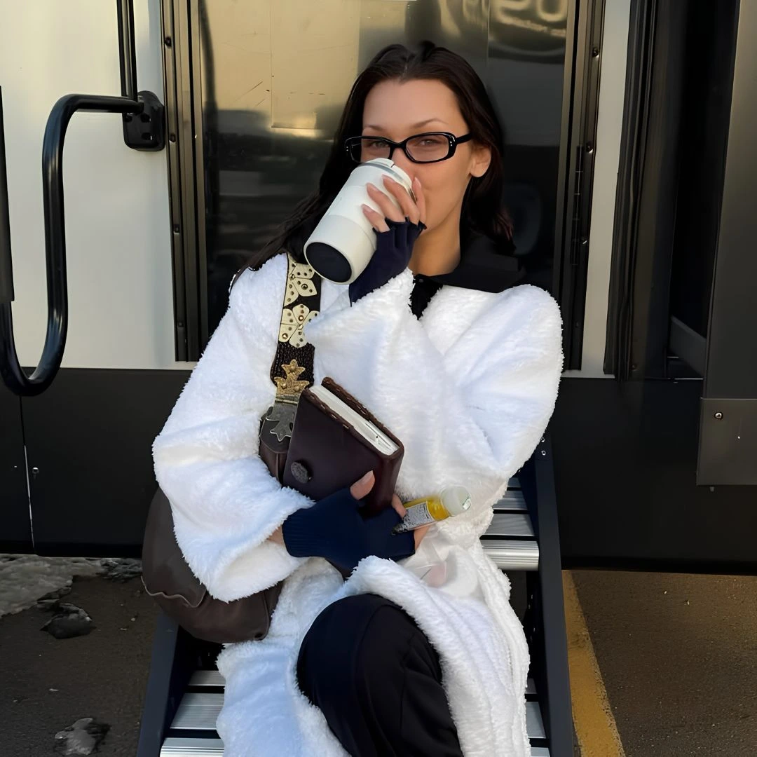 Woman in a white fluffy coat and glasses sits on bus steps, sipping from a to-go cup while holding a wallet and small bottle.