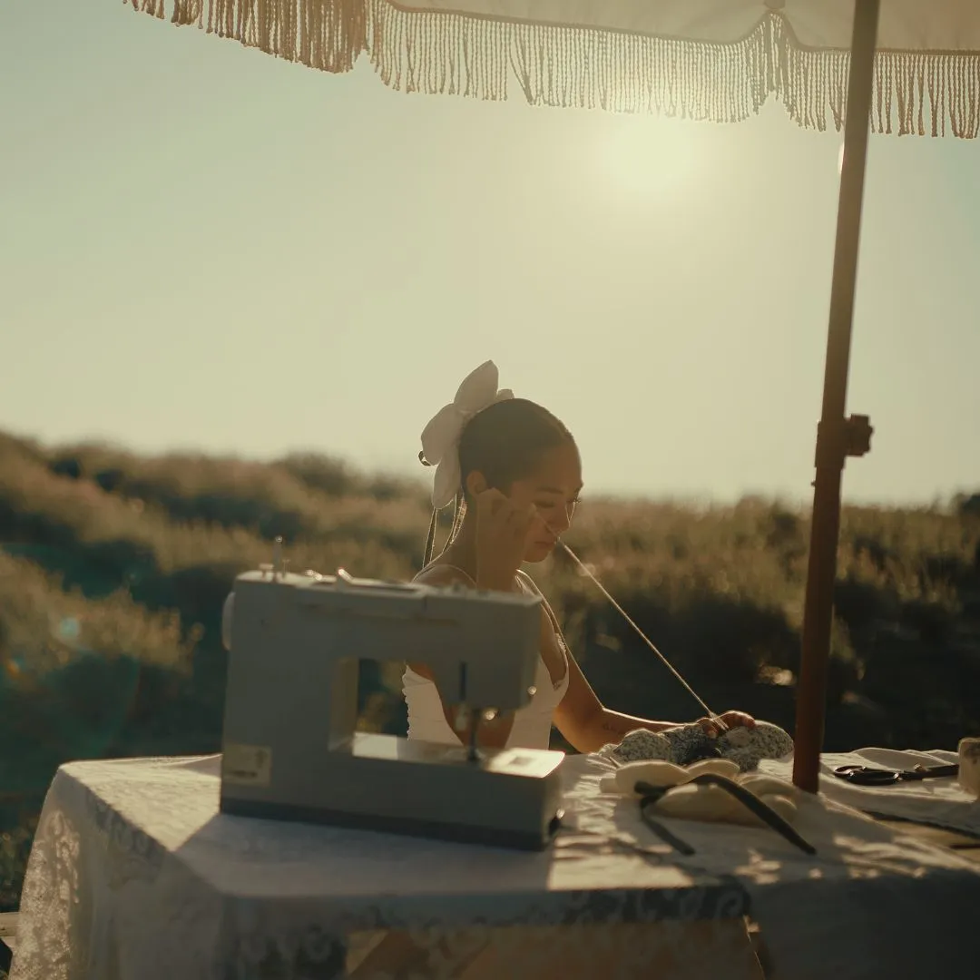 A woman sewing at a table outdoors under a fringed umbrella, talking on a phone as she works with fabric and a sewing machine at sunset.