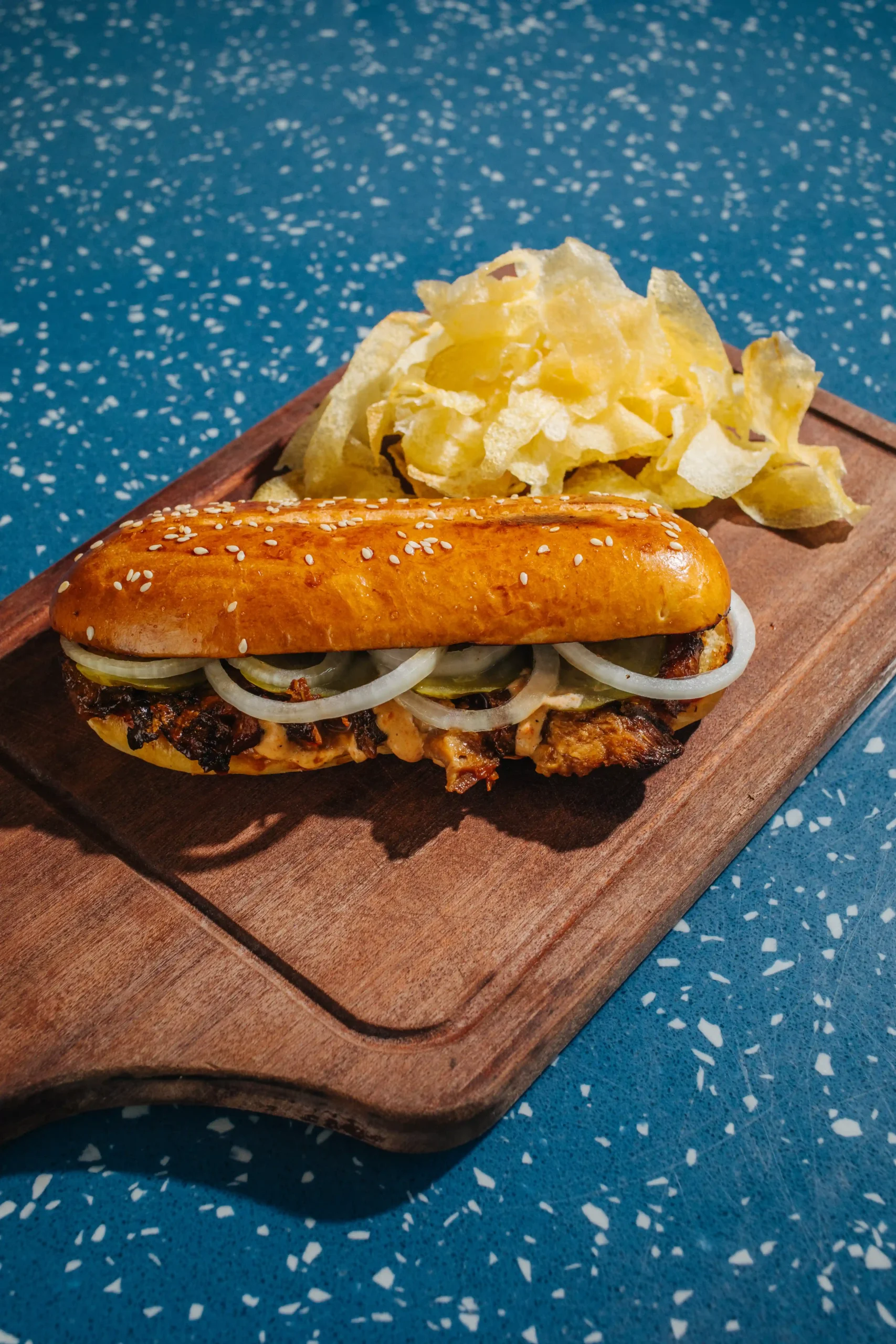 Beef sandwich with onions in a sesame bun on a wooden board, with a side of potato chips in the background.