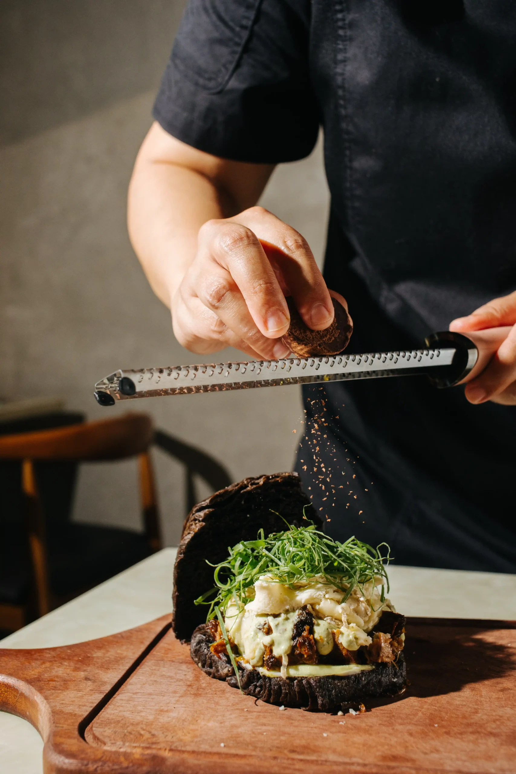 Person in a black shirt grating cheese over an open-faced sandwich with a creamy filling and greens on a wooden board.