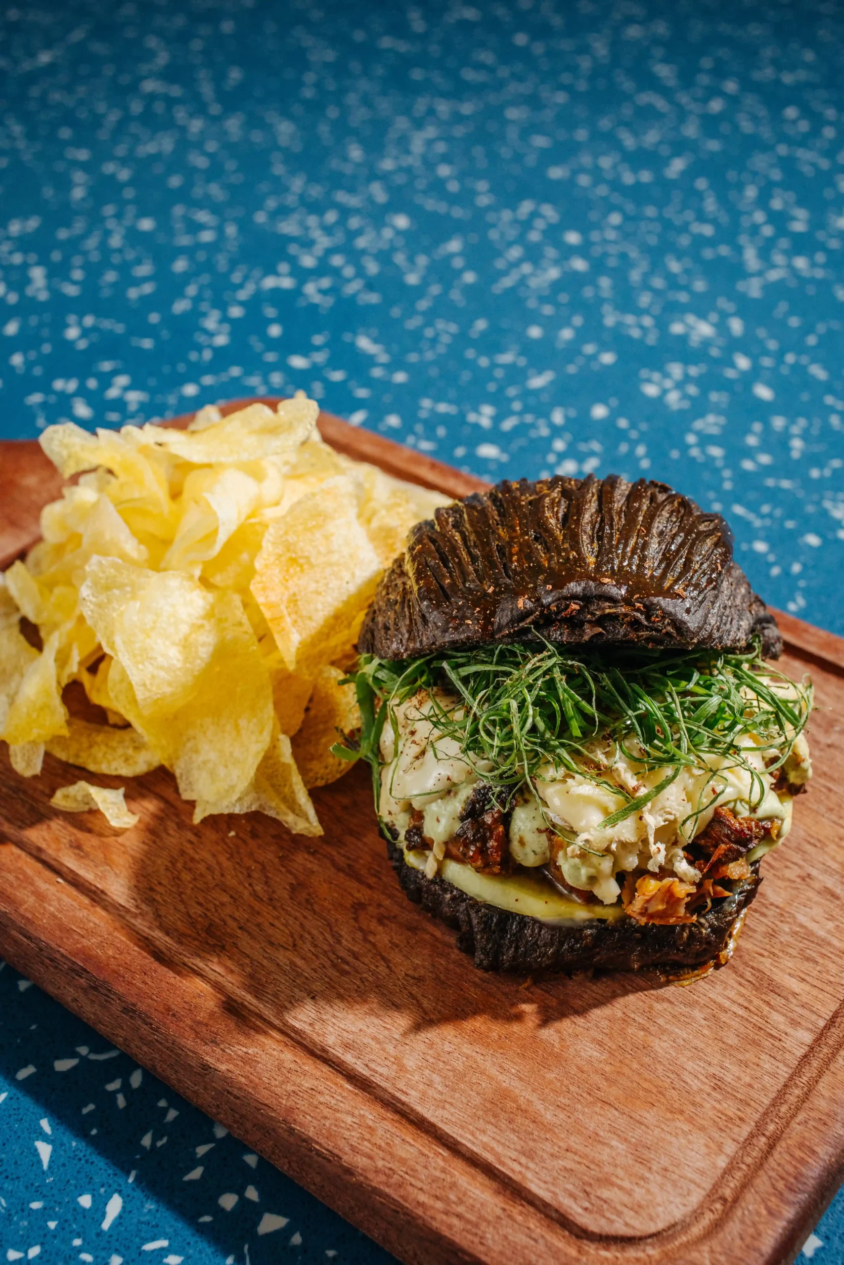 Gourmet burger on a wooden board with a dark bun, greens, and melted filling, beside a pile of crispy potato chips against a blue speckled background