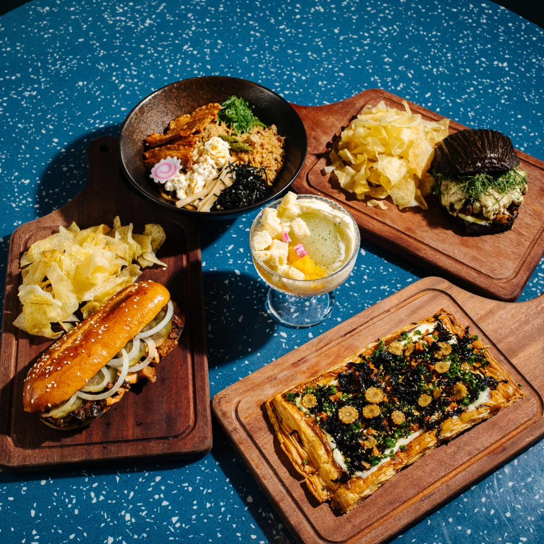 Assorted gourmet dishes arranged on wooden boards: hot dog with onions, a savory tart, a bowl with toppings and greens, and a glass of cocktail on a blue speckled table with potato chips nearby.
