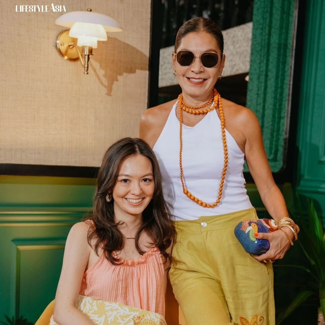 Two women posing together outdoors and smiling; seated woman in a pink top and printed bag, standing woman in a white halter top with yellow pants and orange bead necklace wearing sunglasses.